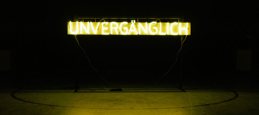 Gelber Neon-Schriftzug mit dem Wort "Unvergänglich" in einem dunklen Raum. Mehr Informationen zum Werk und genauere Beschreibung: https://www.schwabach.de/de/infoseiten-kulturportal/13091-ortung-14-station-13-altes-deutsches-gymnasium-turnhalle.html